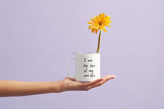 A hand balancing a mug that says "I am the hero of my own life." When we learn how to feel more motivated, we can definitely feel this way.