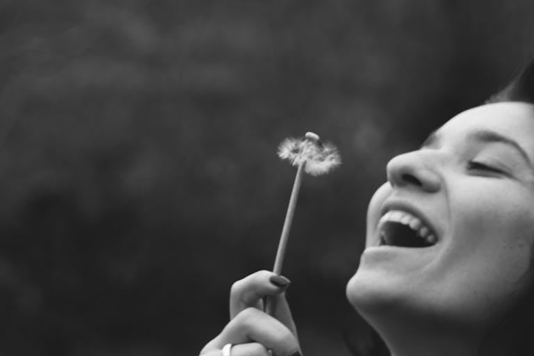 A joyful woman smiling while blowing on a dandelion in a black and white portrait, choosing to be optimistic.