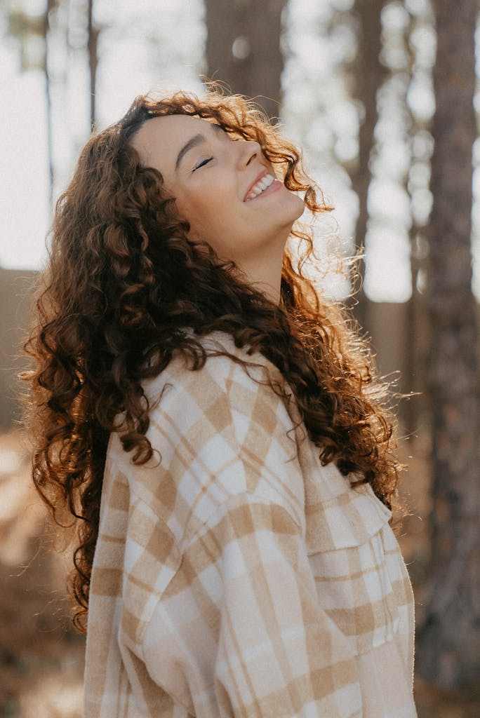 Smiling woman enjoying nature with eyes closed, curly hair shimmering in sunlight.