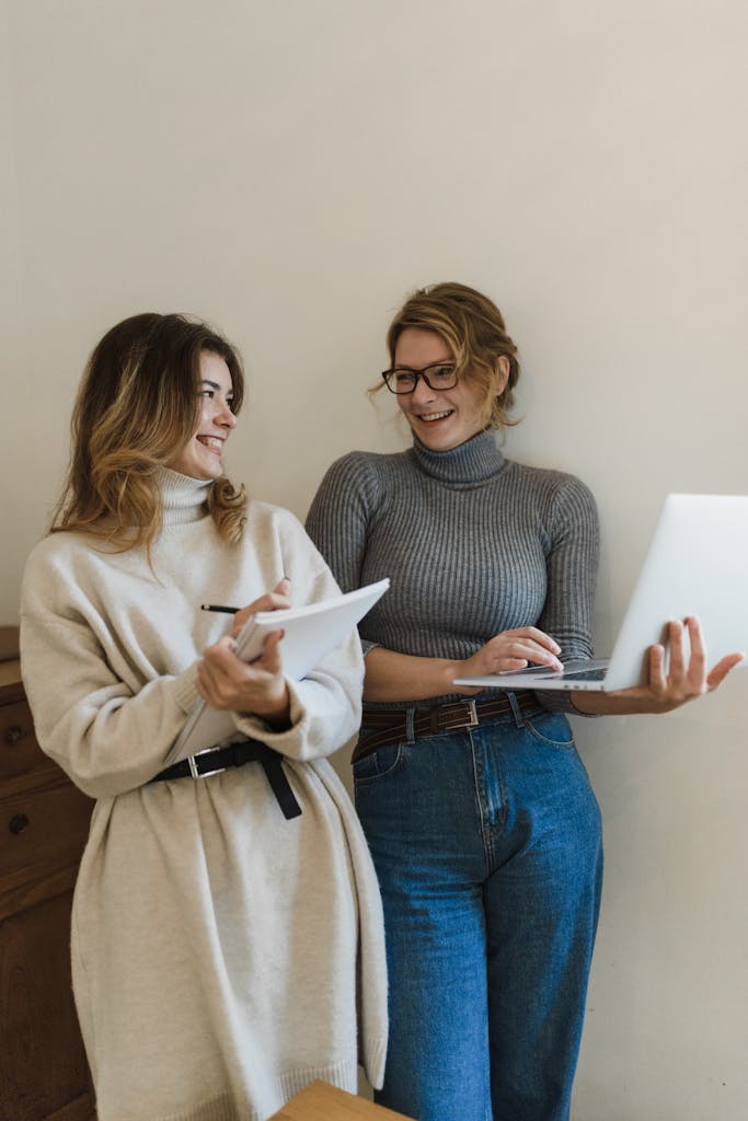 Two women standing indoors, smiling and working with a laptop and notepad.